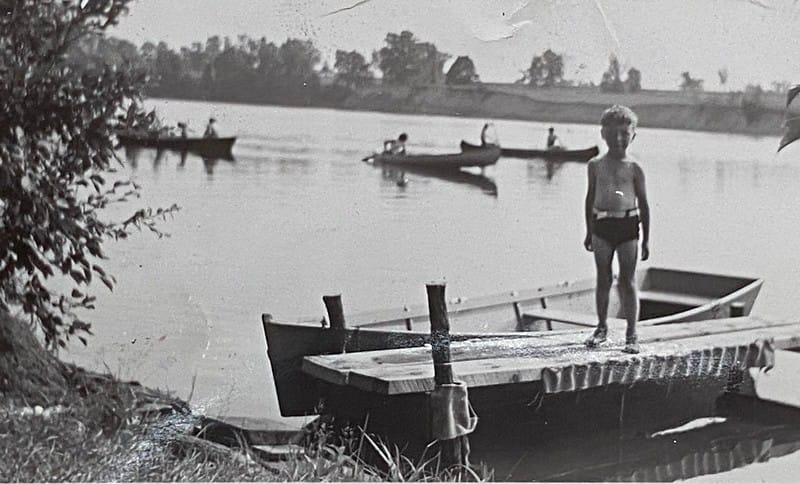 True vintage black & white photo of a boy standing on a dock, circa 1940s. In the background, people row in canoes.